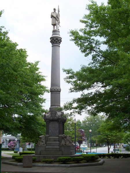 A Civil War Monument shows a soldier standing atop a tall pillar clutching a flag on a pole in one hand while his other hand rests on the hilt of a sword handing at his waist.